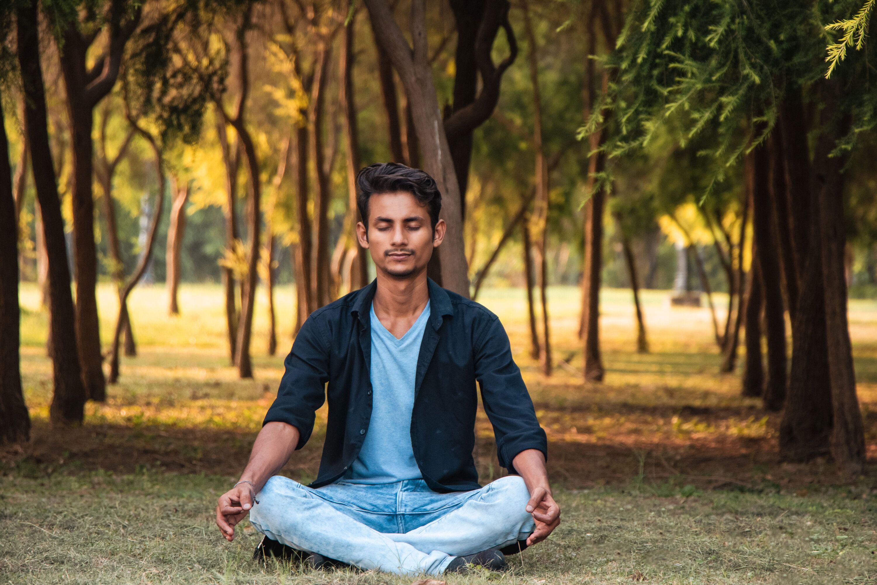 Man sitting cross-legged on the grass in a park with his eyes closed, breathing calmly among trees
