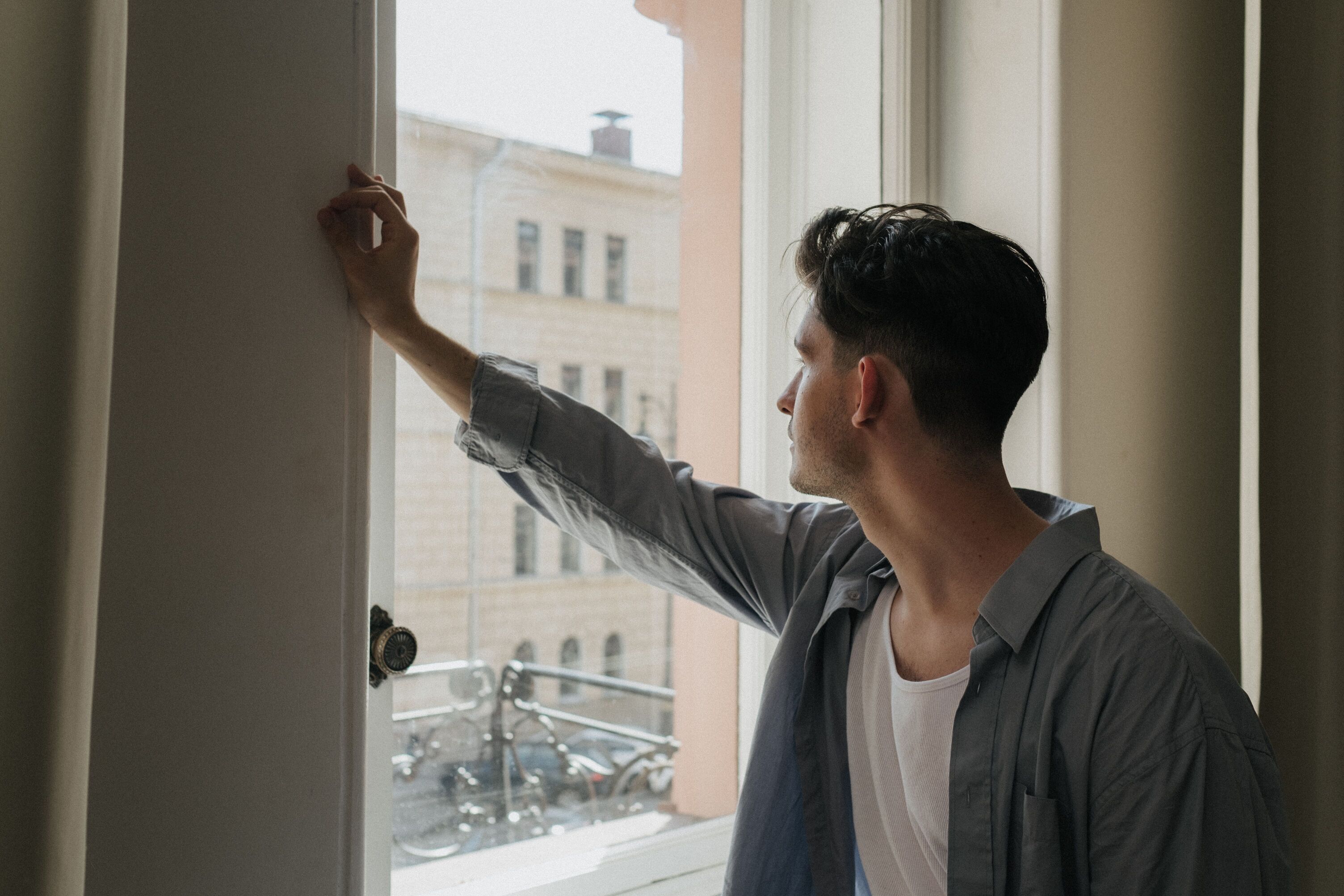 Young man in a grey shirt leaning on a window frame and looking out at city buildings
