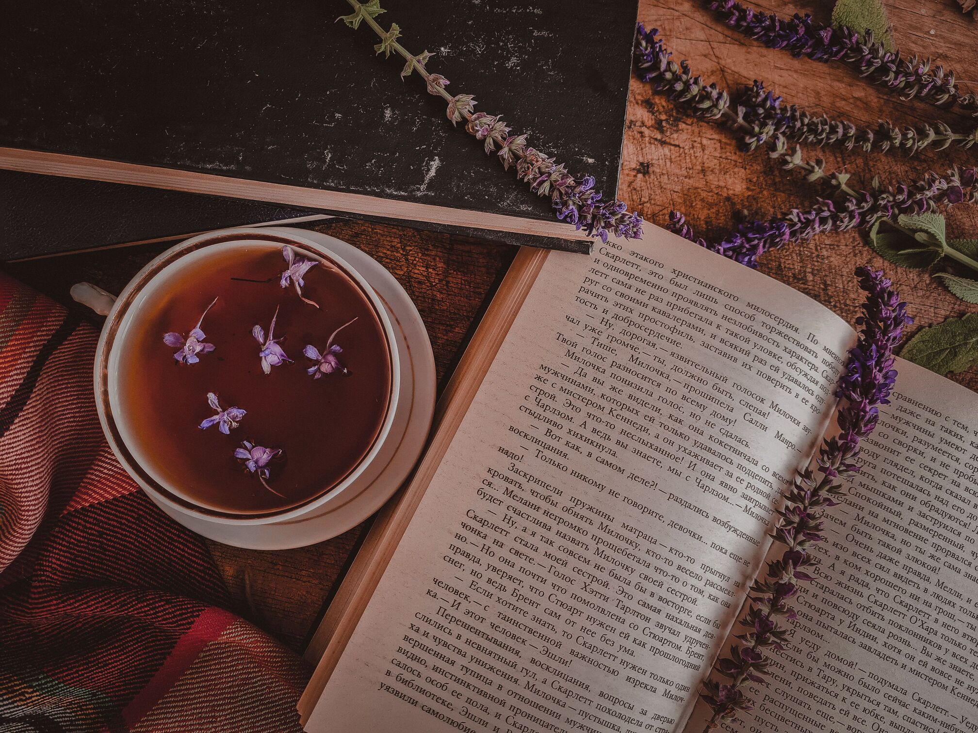 Cup of herbal tea with small purple flowers next to an open book and lavender sprigs on a wooden table