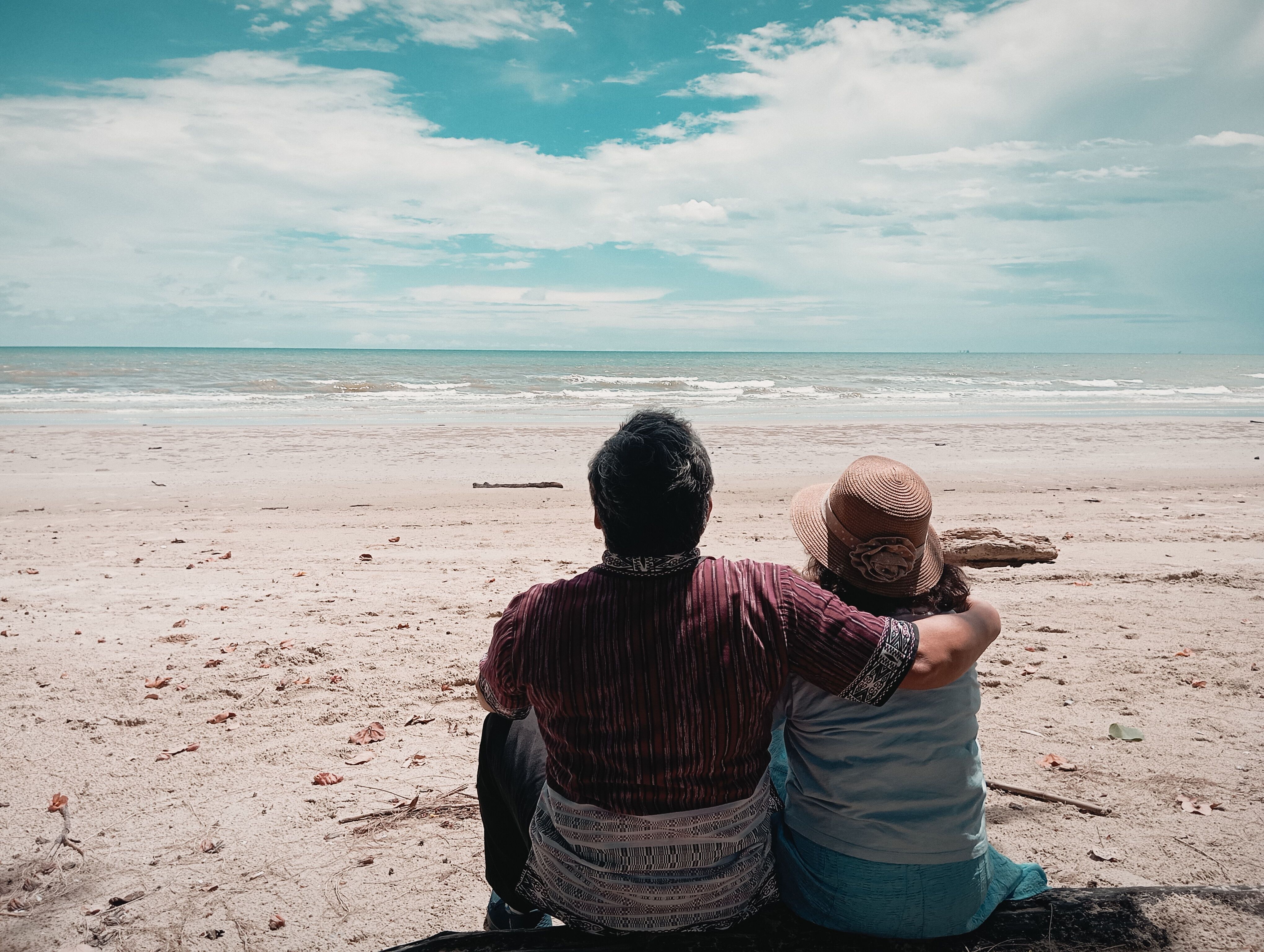 Couple sitting close together on a sandy beach and looking at the sea under a bright cloudy sky
